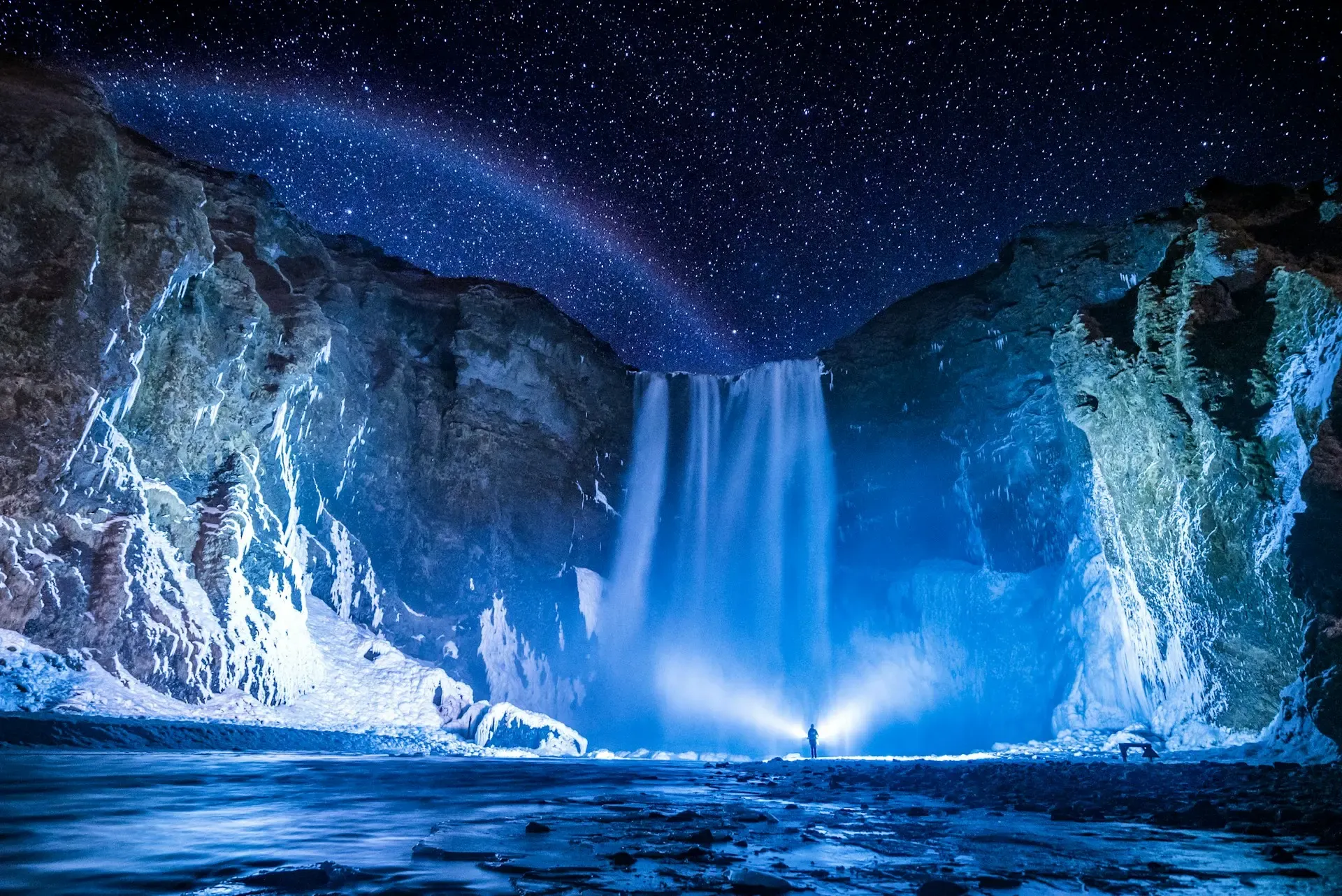 Cascada de Skógafoss iluminada de azul bajo un cielo estrellado, con una persona junto a la base, Islandia