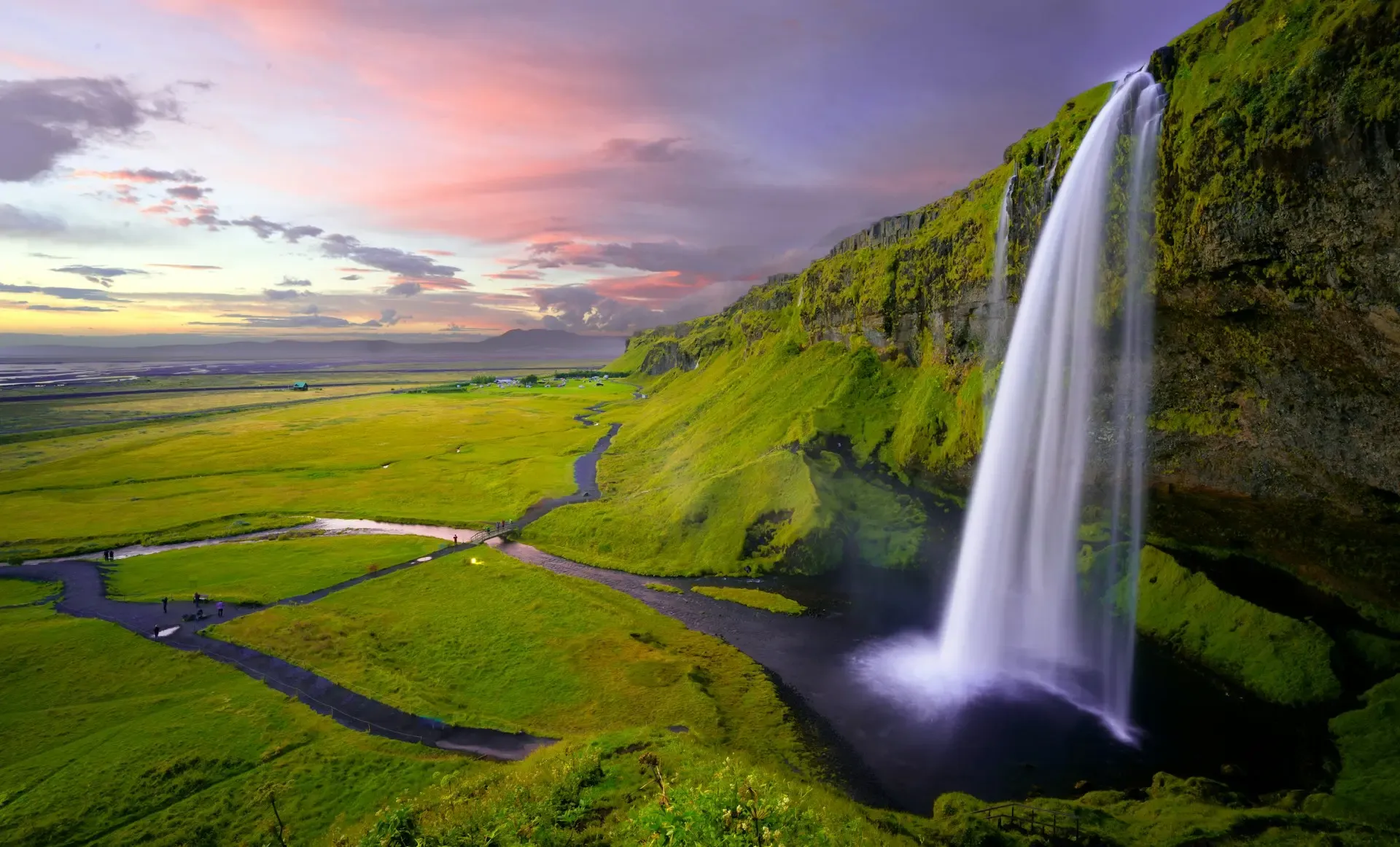 Cascada de Seljalandsfoss cayendo sobre un valle verde al atardecer, con el cielo teñido de rosa, Islandia