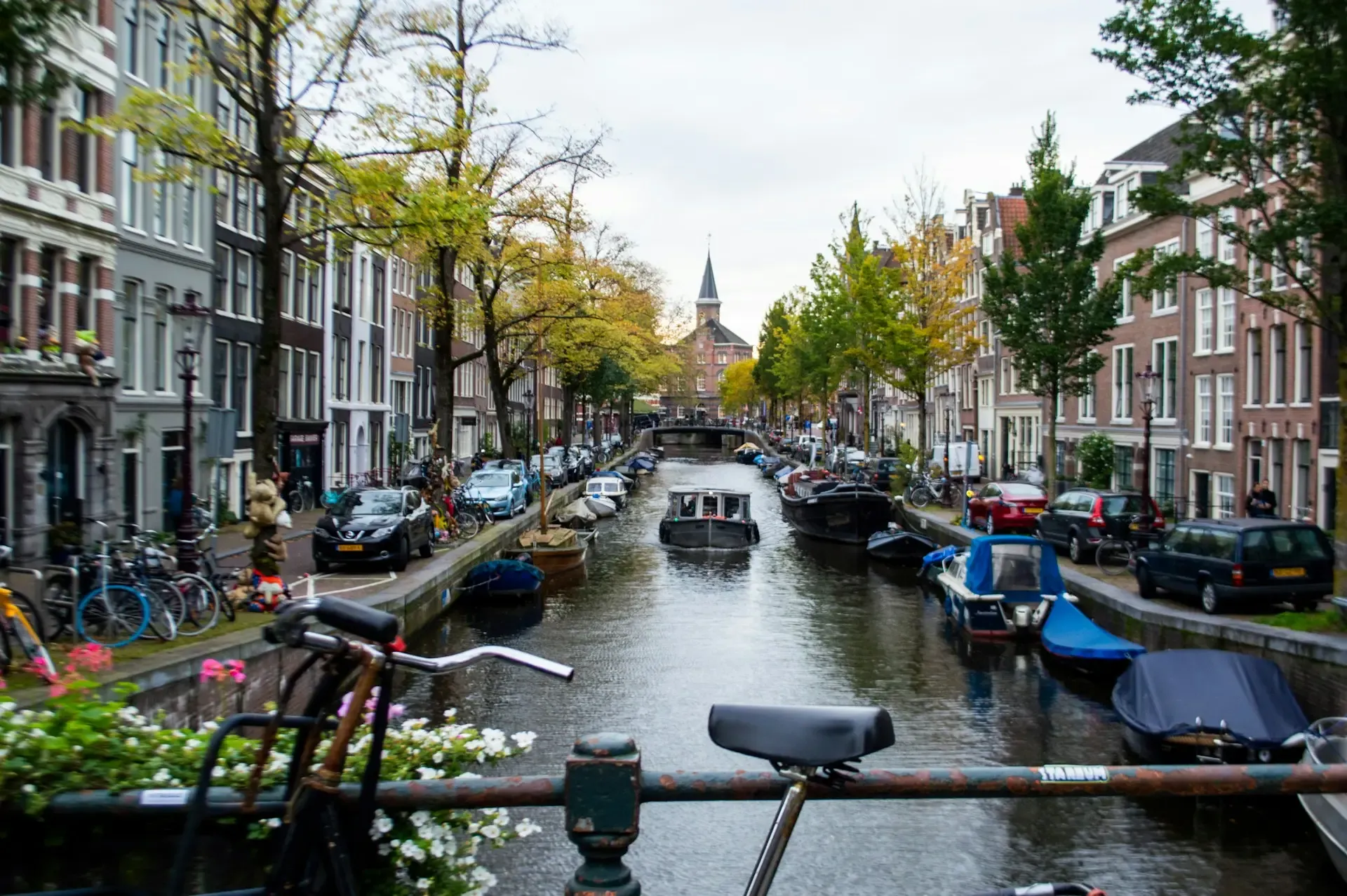 Canal de Ámsterdam flanqueado por casas tradicionales y bicicletas aparcadas, con barcazas amarradas y una torre al fondo