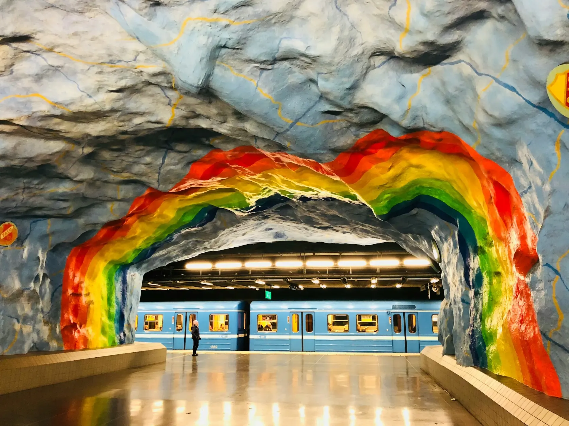 Estación de metro de Estocolmo decorada con un arcoíris pintado sobre roca viva, con un tren azul en el andén