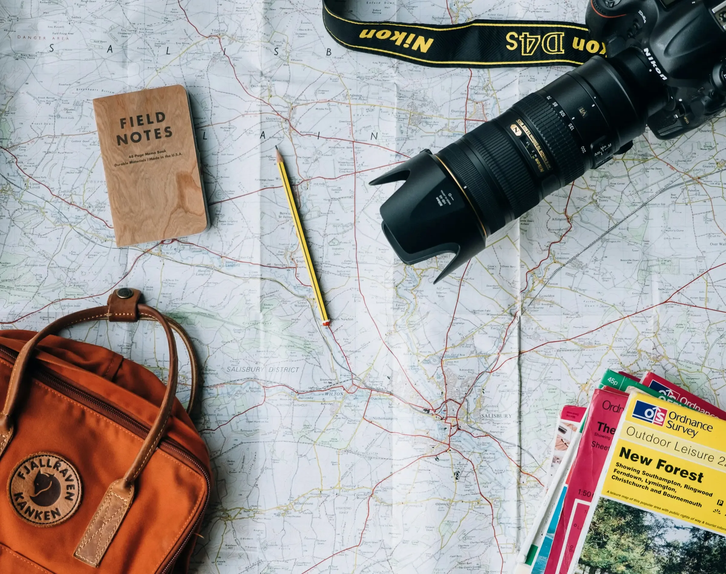 Mapa de viaje desplegado junto a una cámara réflex, una libreta de notas, un lápiz y una mochila, listos para planificar una aventura